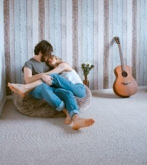 自宅のような場所で男性の膝の上に女性が足をのせてキスしようとしている。その右横にはギターが立てかけられている